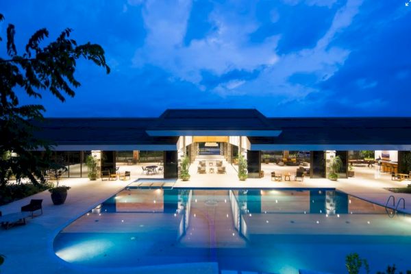 A modern building with large glass openings is illuminated at night, featuring a central swimming pool and surrounded by lounge areas.