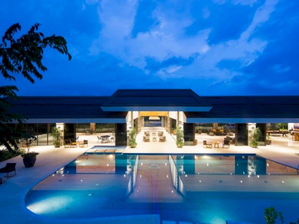 A modern building with large glass openings is illuminated at night, featuring a central swimming pool and surrounded by lounge areas.