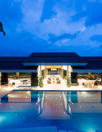 The image shows a luxurious building with a large, illuminated pool at night, surrounded by outdoor seating and lush landscaping.