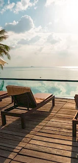 A serene poolside view with lounge chairs, umbrellas, and palm trees facing the ocean under a sunny sky.
