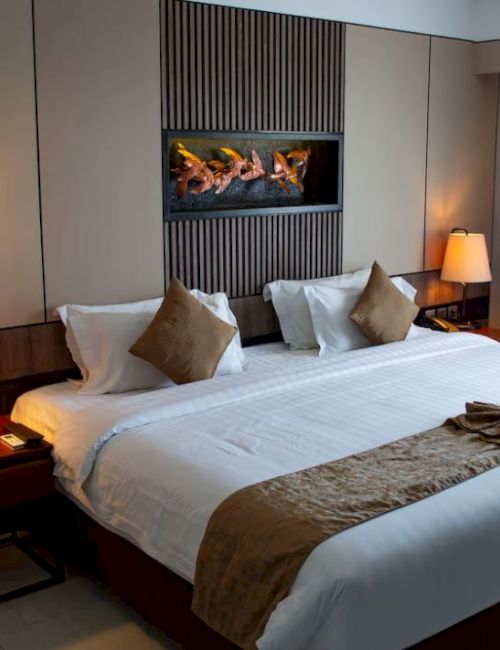 A cozy hotel room with a large bed, decorative pillows, lamps, a wall-mounted fireplace, and curtains beside a window.