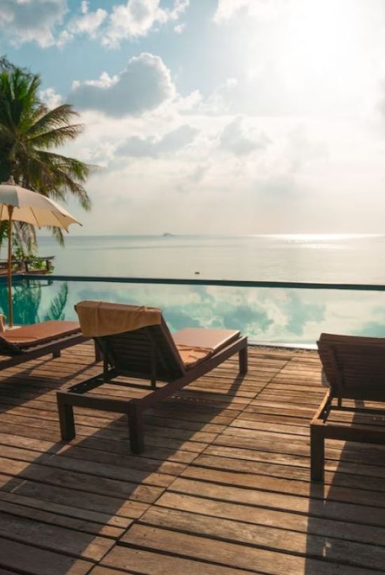 A serene poolside scene with lounge chairs and umbrellas, overlooking a calm ocean under a bright, partly cloudy sky.