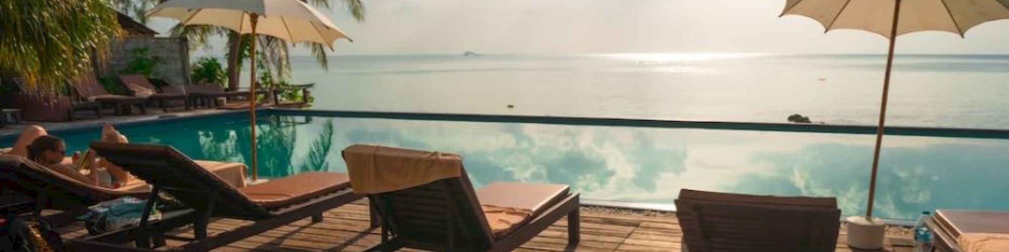 A tranquil poolside scene with deck chairs, umbrellas, palm trees, and a serene water view under a partly cloudy sky.