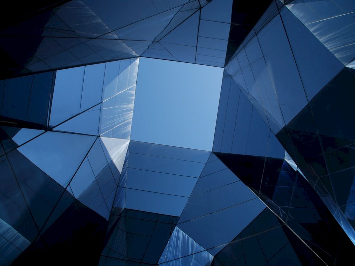 The image shows a geometric architectural structure with reflective glass panels, forming a square opening against a blue sky.