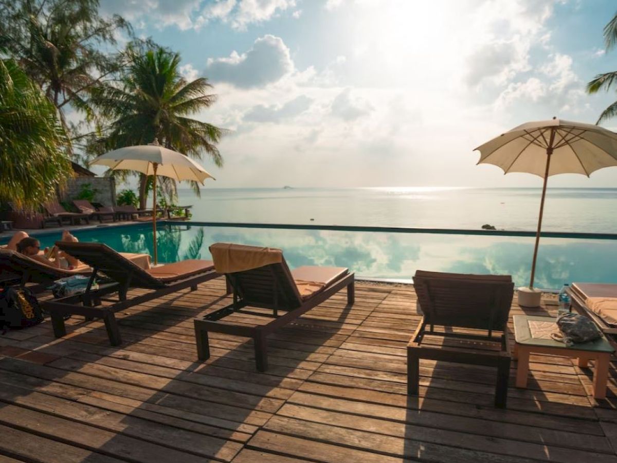 A serene poolside view with lounge chairs, umbrellas, and palm trees overlooking a calm body of water under a bright sky.