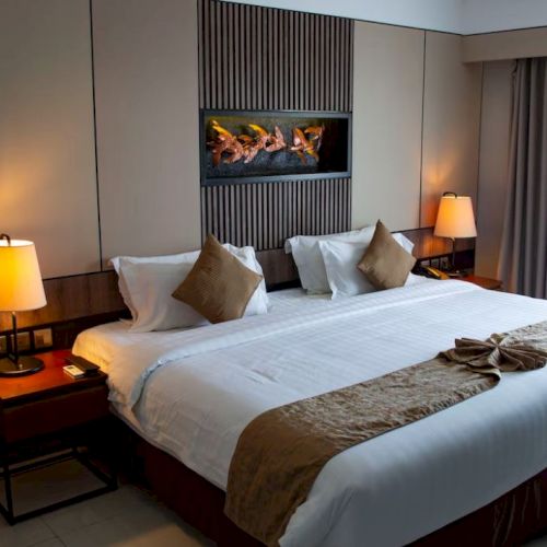 A well-lit hotel room with a large bed, two side lamps, decorative pillows, a tapestry on the wall, and curtains by a window.