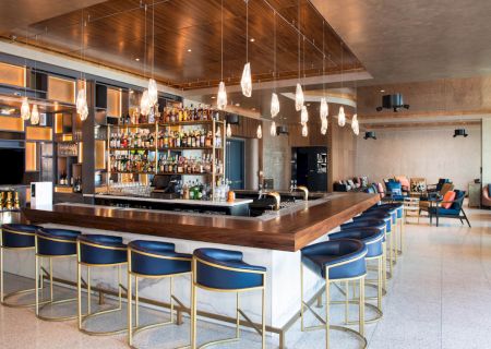 A modern bar with a central counter, blue stools, hanging lights, and a well-stocked shelf. Lounge seating is in the background.