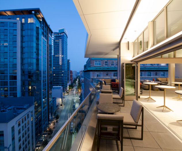 A modern rooftop terrace at dusk with tables and chairs overlooks a cityscape of tall buildings lining a street.