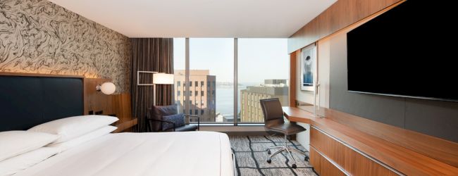 A modern hotel room with a large bed, desk, chair, wide window view, wall-mounted TV, and stylish decor.