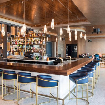 A stylish bar area with a central counter, surrounded by blue chairs, pendant lights, and a well-stocked liquor shelf.