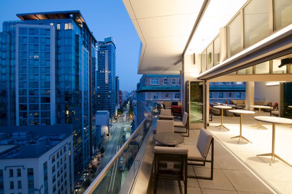 A modern balcony with seating overlooks a cityscape with tall buildings at dusk, creating an urban and tranquil atmosphere.