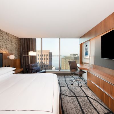 A modern hotel room with a large bed, wall art, a desk, chair, and flat-screen TV, plus a city view through a large window.