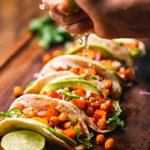 A hand squeezes lime over a row of vibrant tacos filled with chickpeas, avocado, and veggies on a wooden board.