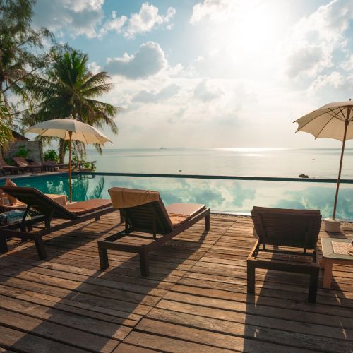 A serene beach scene with lounge chairs, umbrellas, and an infinity pool overlooking the ocean during a sunny day with palm trees.