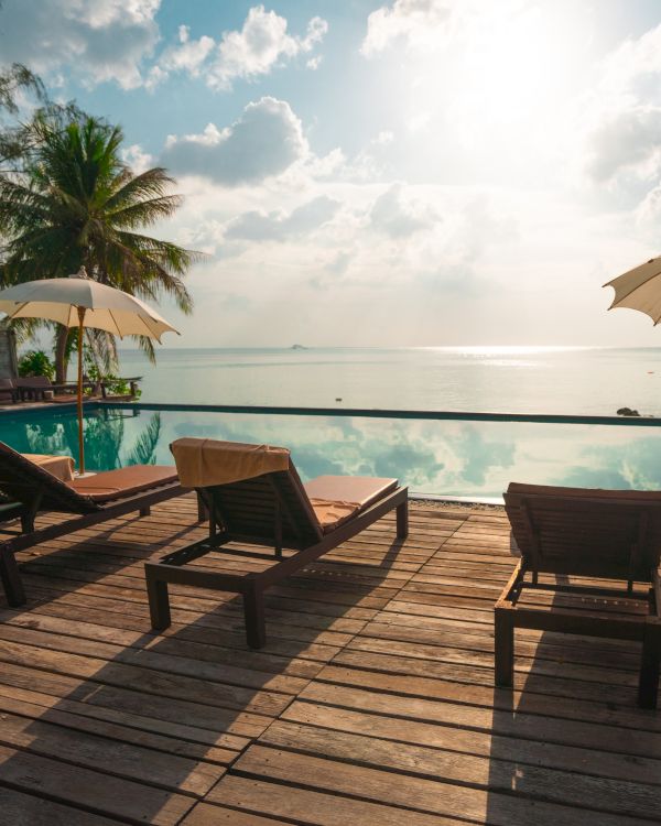 A serene beach scene with lounge chairs, umbrellas, and an infinity pool overlooking the ocean during a sunny day with palm trees.