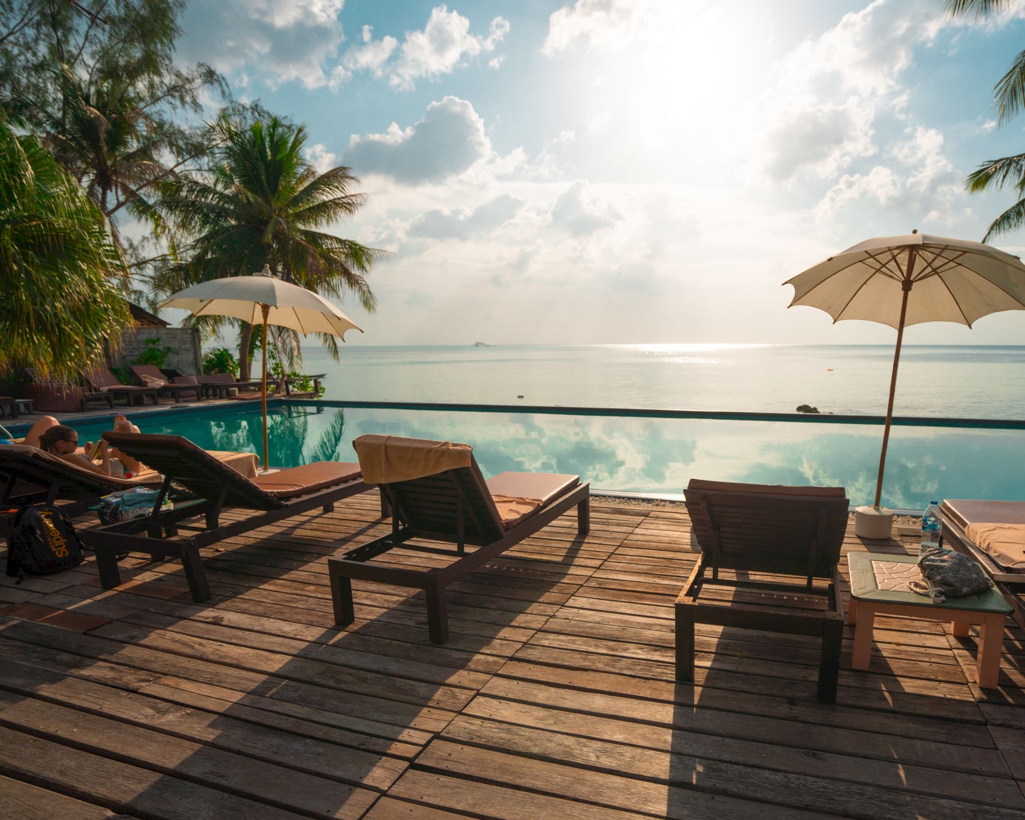 Poolside loungers and umbrellas by an infinity pool overlooking a calm sea, with palm trees and a cloudy sky in the background.