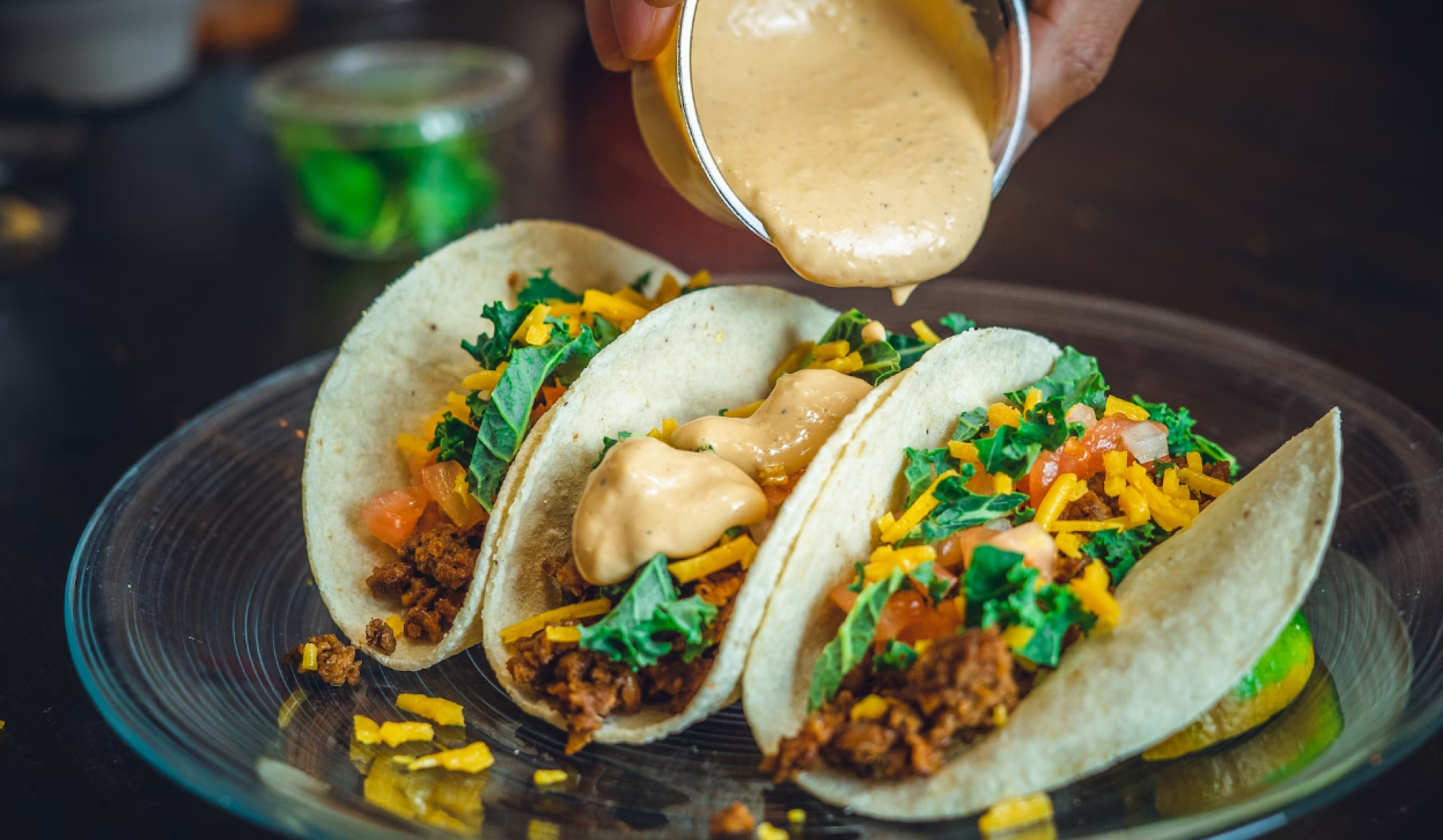 Three tacos filled with beef, cheese, lettuce, and tomatoes are being topped with a creamy sauce on a glass plate.