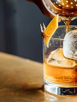 A cocktail is being poured into a glass with ice and a garnish on a metallic surface, finishing it with a strainer.