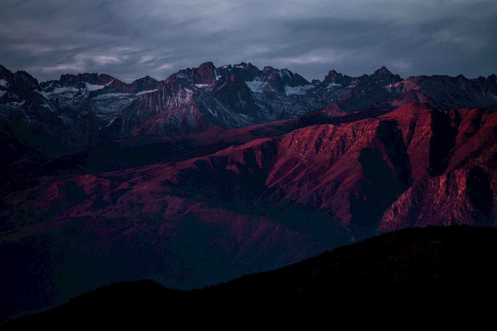 A mountain range at dusk, with rugged peaks and slopes bathed in red light under a cloudy sky.