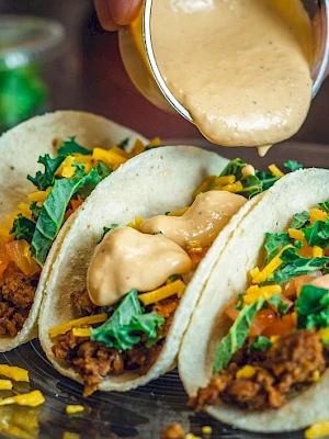 Three tacos filled with ground meat, shredded cheese, lettuce, and tomato on a plate; sauce is being poured on top.