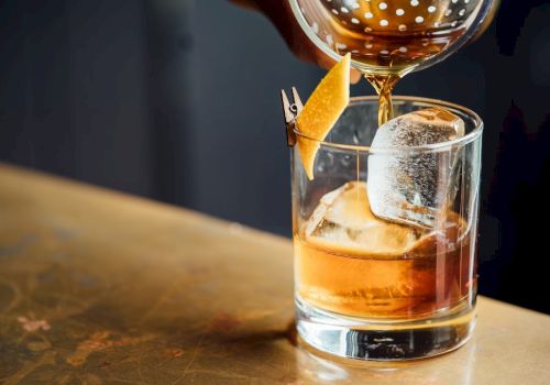 A cocktail being poured over ice in a glass, garnished with a twist of citrus peel.