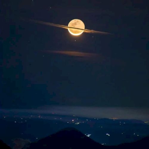 The image shows a bright moon with clouds resembling Saturn's rings, over a dark landscape with distant lights and silhouetted hills.