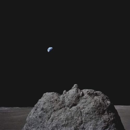 A rocky surface with a large boulder in the foreground and Earth visible in the distant, dark sky.