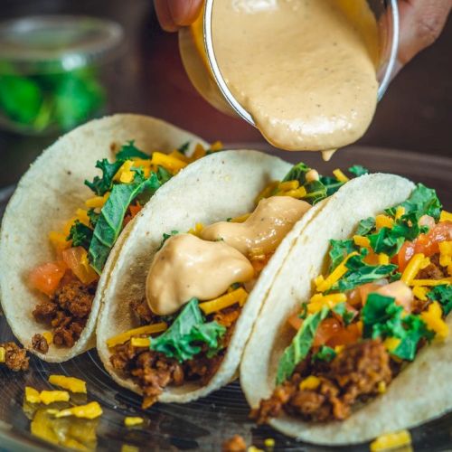 Three tacos with seasoned meat, cheese, lettuce, and tomatoes, being topped with creamy sauce on a plate.