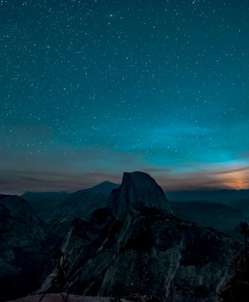 A starry night sky over a silhouetted mountainous landscape, with a glowing horizon and scattered clouds enhancing the scene.