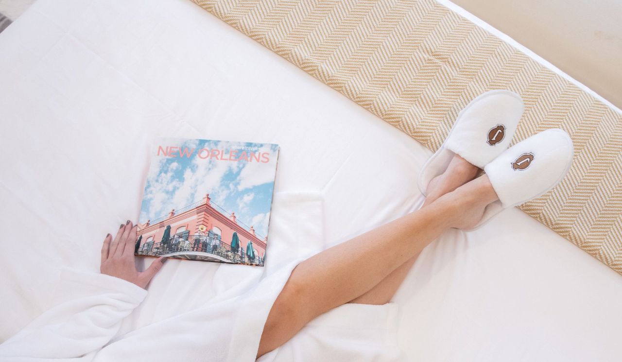 A person's legs and feet in white slippers on a bed with a magazine.