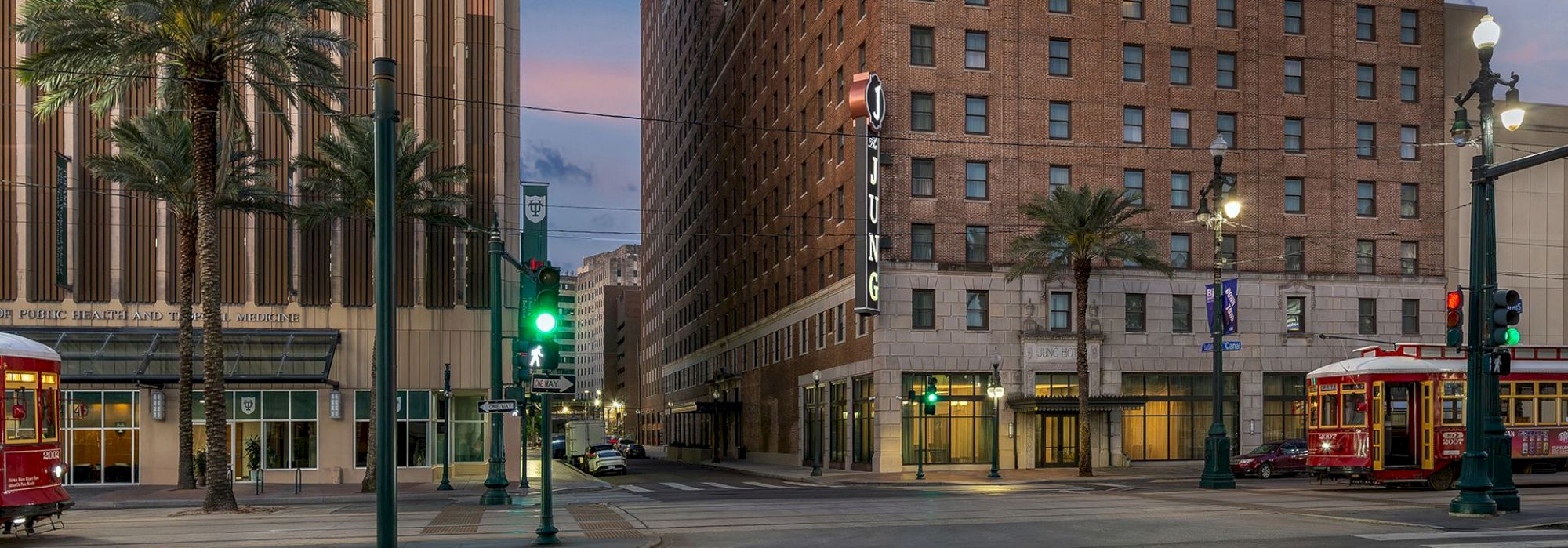 Street with tall buildings, palm trees, traffic lights, and two red streetcars at dusk.