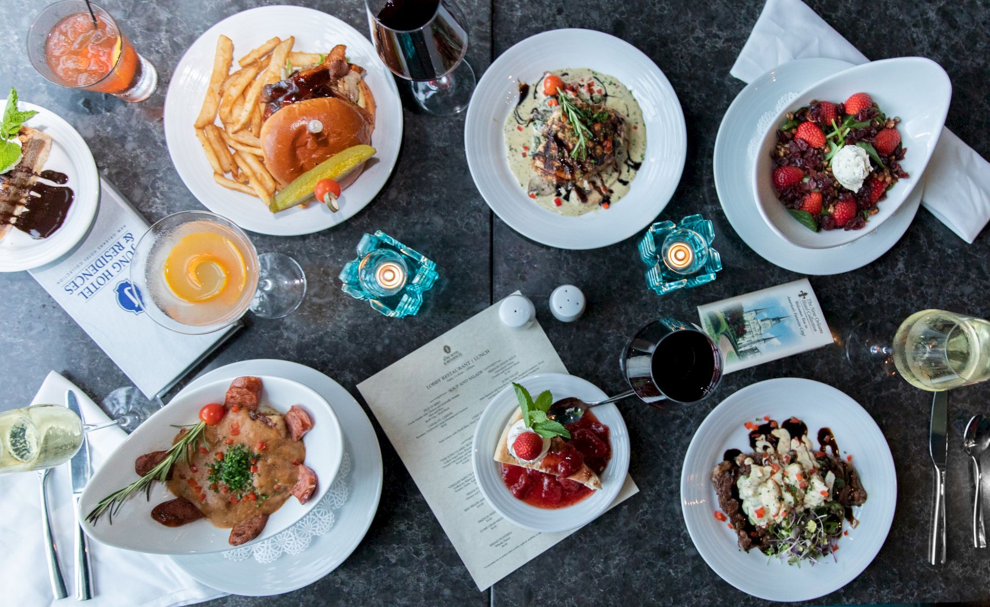 An overhead view of a table with various dishes and drinks, featuring plates of food, wine glasses, and a menu in the center.
