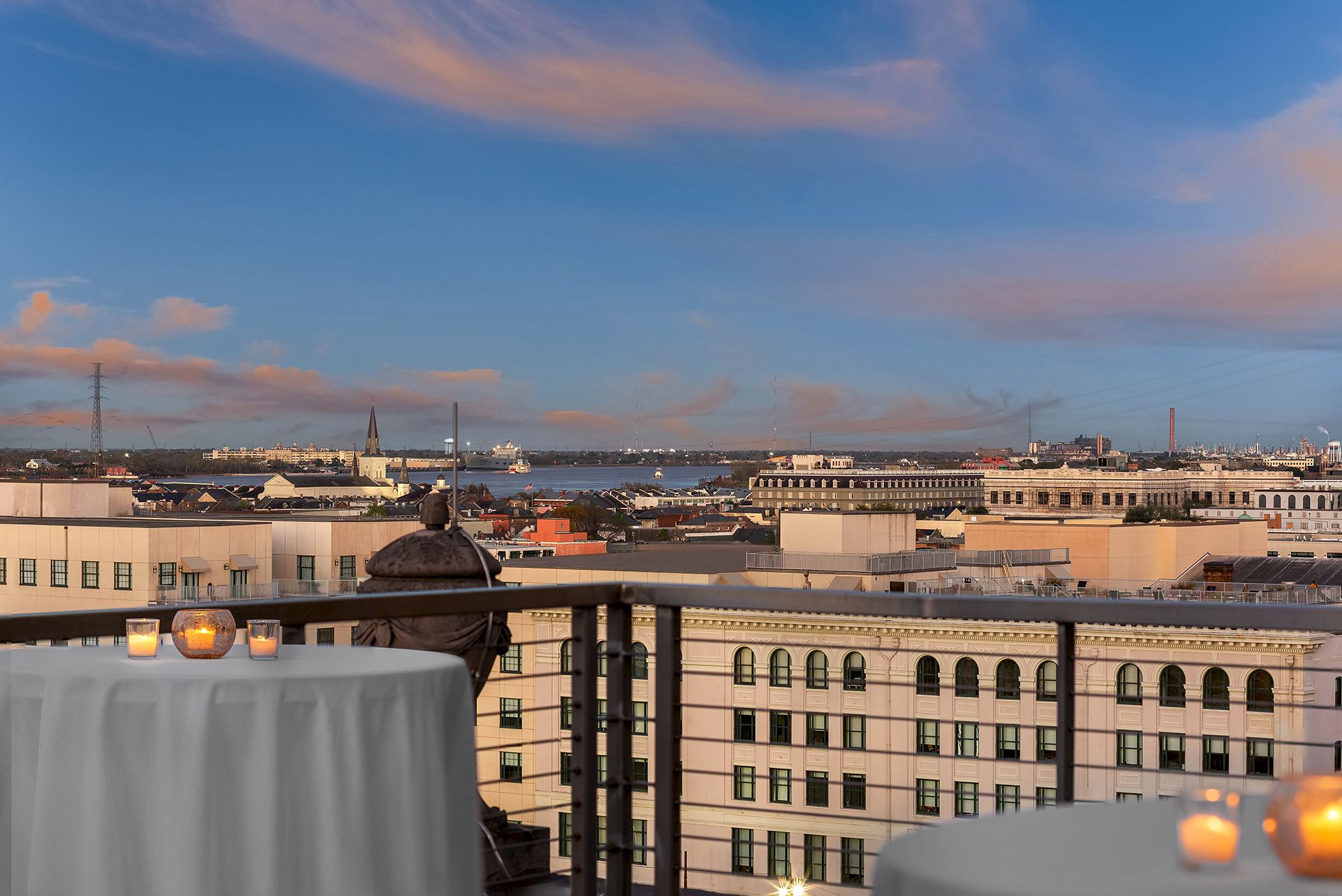 A rooftop view overlooks a cityscape, featuring buildings, a river, and a sky at sunset, with tables adorned with candles in the foreground.