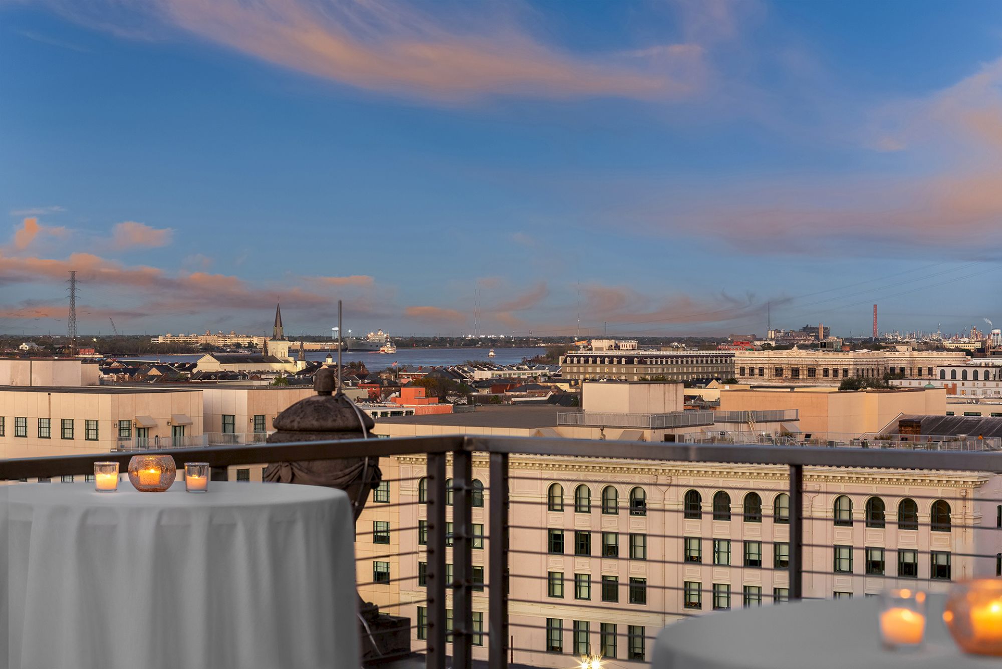 A rooftop view overlooks a cityscape, featuring buildings, a river, and a sky at sunset, with tables adorned with candles in the foreground.