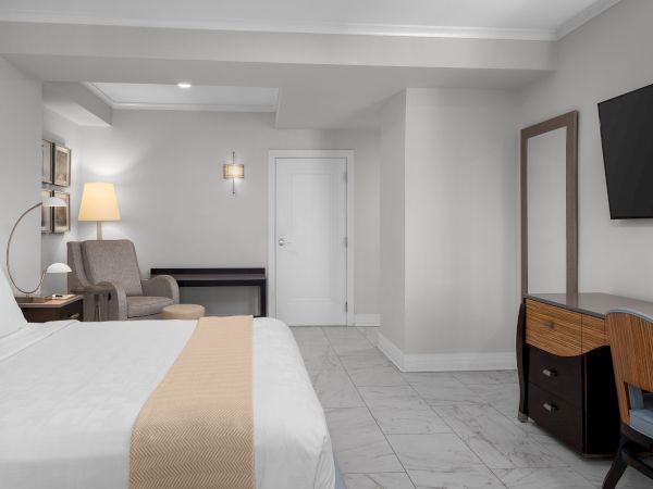 The image shows a modern hotel room with a bed, armchair, lamp, TV, desk, and chair on a tiled floor.