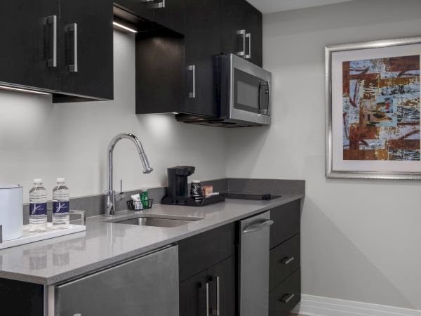 A modern kitchenette with a sink, microwave, coffee maker, two water bottles, and abstract art on the wall.