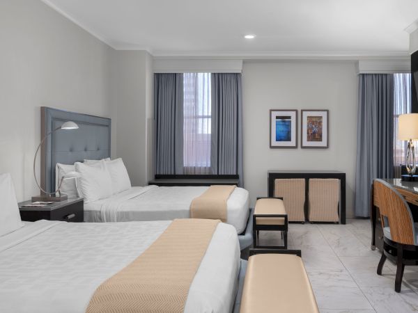The image shows a modern hotel room with two beds, a desk, and two chairs, featuring neutral colors and framed artwork on the wall.