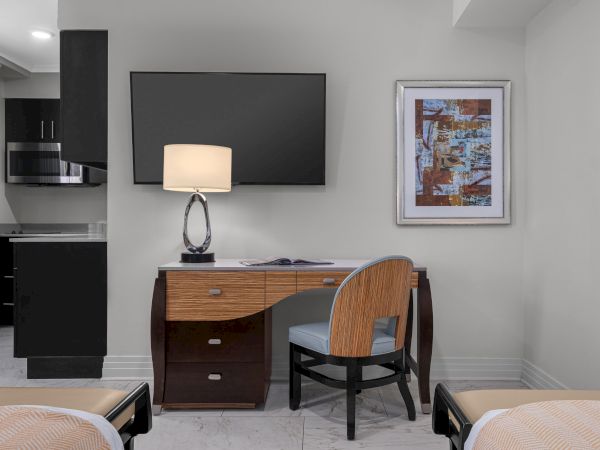 A hotel room with a desk, chair, lamp, and TV. There's abstract art on the wall. Kitchen area with microwave visible.