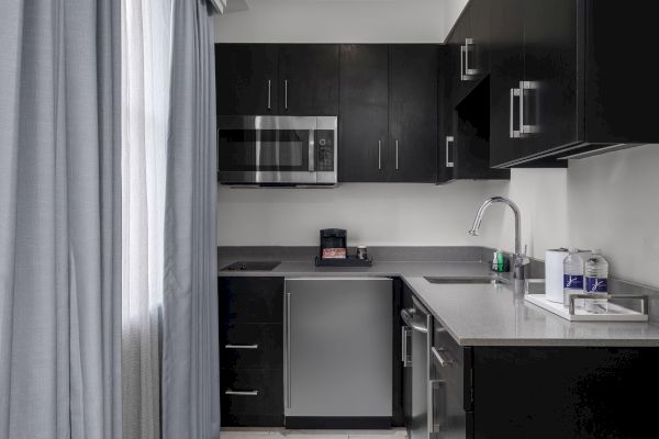 A small kitchen with dark cabinets, a microwave, fridge, coffee maker, sink, and water bottles on the counter.