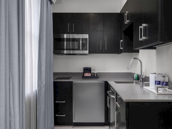 A small kitchen with dark cabinets, a microwave, fridge, coffee maker, sink, and water bottles on the counter.