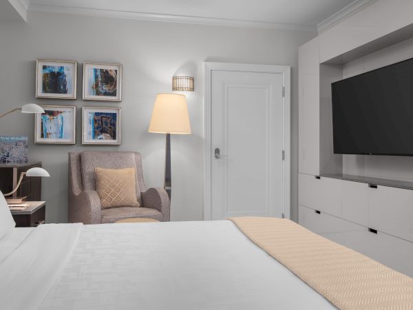 A cozy room featuring a bed, armchair, four wall artworks, a floor lamp, and a TV. A "New Orleans" book sits on the side table.