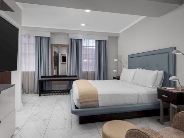 A modern bedroom with a large bed, side tables, lamps, a mirror, blue curtains, and a wall-mounted TV, featuring marble floors.