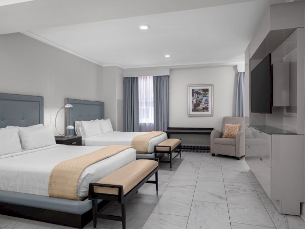 A modern hotel room with two beds, a TV, armchair, benches, and a window with curtains.