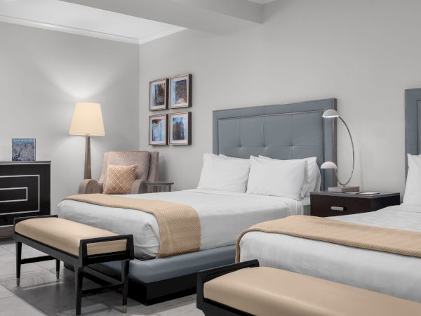 The image shows a hotel room with two beds, a chair, a lamp, and framed pictures on the wall.