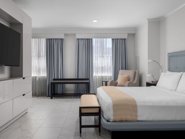 A modern hotel room with a bed, armchair, large TV, and curtains over the windows.