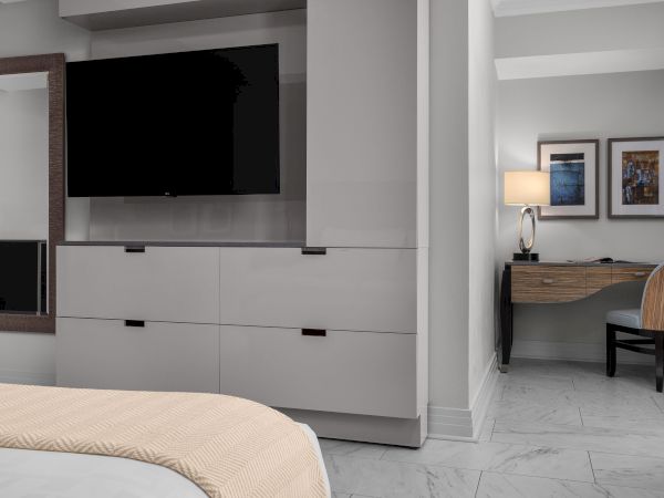 A modern room features a large TV, dresser, mirror, bed with beige blanket, desk with lamp, chair, and framed artwork on the wall.