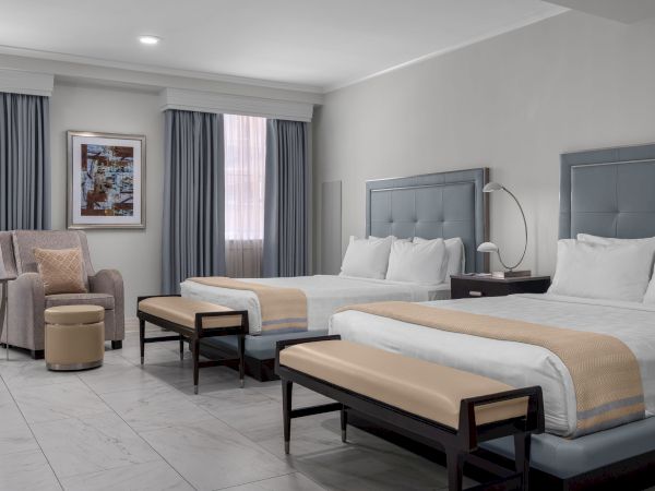 The image shows a hotel room with two double beds, a chair, an ottoman, a side table, and a framed artwork on the wall.