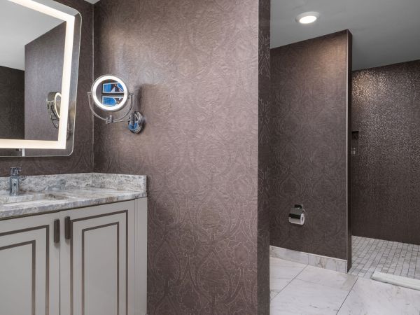 The image shows a modern bathroom with a marble countertop, lit mirror, wall-mounted magnifying mirror, and a walk-in shower.