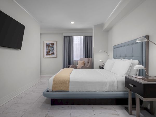 The image shows a modern bedroom with a bed, bedside lamps, an armchair, a framed picture, and a wall-mounted TV.