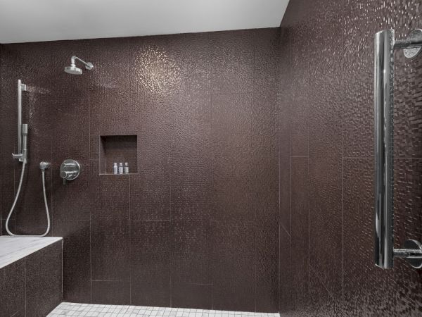 A modern shower with a bench, dark textured walls, a rain showerhead, handheld shower, and small bottles in a wall niche.